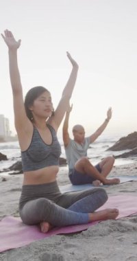 Çift ırklı mutlu çiftin yoga yaptığı ve kumsalda yavaş çekimde meditasyon yaptığı dikey bir video. Kaliteli zaman, yaşam tarzı ve tatil konsepti harcamak.