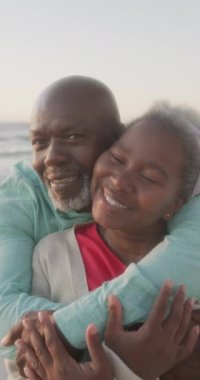 Yaşlı Afro-Amerikan çiftin kumsalda yavaşça kucaklaşmalarını gösteren dikey bir video. Kaliteli zaman, yaşam tarzı, emeklilik ve tatil konsepti.