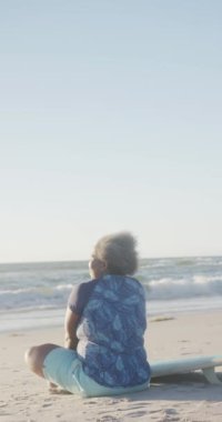 Ağır çekimde kumsalda oturan mutlu Afrikalı Amerikalı kadının dikey videosu. Kaliteli zaman, yaşam tarzı, emeklilik ve tatil konsepti.