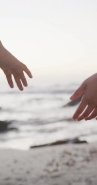 Çift ırklı çiftin kumsalda ağır çekimde el ele tutuşmalarını gösteren dikey bir video. Kaliteli zaman, yaşam tarzı ve tatil konsepti harcamak.