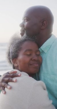 Yaşlı Afro-Amerikan çiftin kumsalda yavaşça kucaklaşmalarını gösteren dikey bir video. Kaliteli zaman, yaşam tarzı, emeklilik ve tatil konsepti.