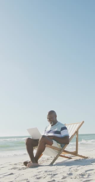 Pionowe Wideo Starszego Afroamerykanina Używającego Laptopa Leżaku Plaży Zwolnionym Tempie — Wideo stockowe