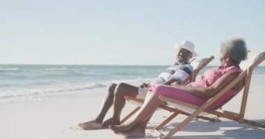 Mutlu Afro-Amerikan çift güverte sandalyelerinde ve sahilde konuşuyorlar, fotokopi uzayı, ağır çekimde. Kaliteli zaman geçirmek, yaşam tarzı, emeklilik ve tatil kavramı.