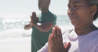 Mutlu Afrika kökenli Amerikalı çift yavaş çekimde yoga yapıyor ve plajda meditasyon yapıyor. Kaliteli zaman geçirmek, yaşam tarzı, emeklilik ve tatil kavramı.