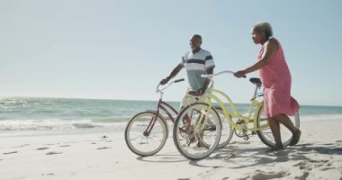 Mutlu son sınıf Afro-Amerikan çifti yavaş çekimde sahilde bisikletleriyle yürüyor. Kaliteli zaman geçirmek, yaşam tarzı, emeklilik ve tatil kavramı.
