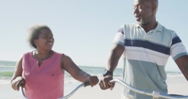Mutlu son sınıf Afro-Amerikan çifti yavaş çekimde sahilde bisikletleriyle yürüyor. Kaliteli zaman geçirmek, yaşam tarzı, emeklilik ve tatil kavramı.