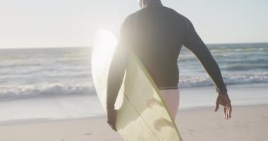 Sahilde yürüyen ve sörf tahtası tutan kıdemli Afro-Amerikan adam. Ağır çekimde. Kaliteli zaman geçirmek, yaşam tarzı, emeklilik ve tatil kavramı.