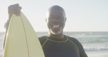 Sahilde sörf tahtası tutan son sınıf Afro-Amerikan adamının portresi. Ağır çekimde. Kaliteli zaman geçirmek, yaşam tarzı, emeklilik ve tatil kavramı.