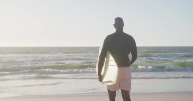 Sahilde yürüyen ve sörf tahtası tutan kıdemli Afro-Amerikan adam. Ağır çekimde. Kaliteli zaman geçirmek, yaşam tarzı, emeklilik ve tatil kavramı.