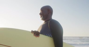 Sahilde yürüyen ve sörf tahtası tutan kıdemli Afro-Amerikan adam. Ağır çekimde. Kaliteli zaman geçirmek, yaşam tarzı, emeklilik ve tatil kavramı.