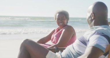 Mutlu Afrika kökenli Amerikalı çift güverte sandalyelerinde oturup kumsalda ağır çekimde konuşuyor. Kaliteli zaman geçirmek, yaşam tarzı, emeklilik ve tatil kavramı.