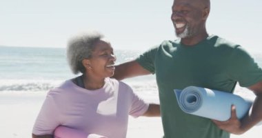 Sahilde yoga minderlerini kucaklayan ve ağır çekimde tutan mutlu Afro-Amerikan çifti. Kaliteli zaman geçirmek, yaşam tarzı, emeklilik ve tatil kavramı.