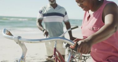 Mutlu son sınıf Afro-Amerikan çifti yavaş çekimde sahilde bisikletleriyle yürüyor. Kaliteli zaman geçirmek, yaşam tarzı, emeklilik ve tatil kavramı.