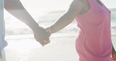 Kıdemli Afro-Amerikan çiftin orta kesiminde kumsalda ağır çekimde el ele tutuşuyorlar. Kaliteli zaman geçirmek, yaşam tarzı, emeklilik ve tatil kavramı.