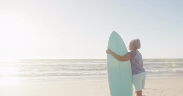 快乐的资深非洲裔美国女人在海滩上抱着冲浪板 动作缓慢 有复制空间 花费高质量的时间 生活方式 退休和度假概念 — 图库视频影像