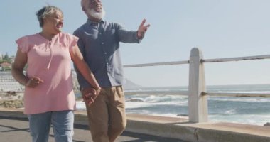 Mutlu kıdemli Afro-Amerikan çifti deniz kenarında ağır çekimde geziniyor. Kaliteli zaman, yaşam tarzı, tatil ve emeklilik.