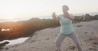 Kıdemli Afro-Amerikan kadın yoga yapıyor ve plajda geriniyor, ağır çekimde. Kaliteli zaman, yaşam tarzı, tatil ve emeklilik.