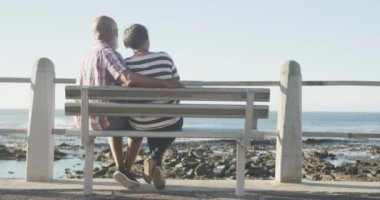 Mutlu son sınıf Afro-Amerikan çifti deniz kenarındaki bankta oturuyor, ağır çekimde. Kaliteli zaman, yaşam tarzı, tatil ve emeklilik.