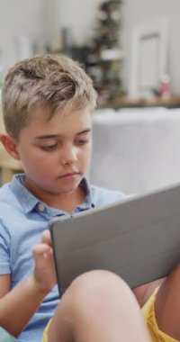 Kanepeye uzanmış ve tablet kullanan beyaz bir çocuğun dikey videosu, ağır çekim. Kaliteli zaman geçirme, ev hayatı ve çocukluk kavramı.