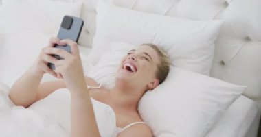 Kafkasyalı mutlu, iri yarı bir kadın. Ağır çekimde yatakta gülüyor ve akıllı telefon kullanıyor. Yaşam tarzı, evde kaliteli zaman geçirme ve vücut kapsamı.