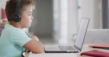 Kafkasyalı mutlu çocuk masada oturuyor ve bilgisayarını online ders olarak kullanıyor, ağır çekimde. Kaliteli zaman geçirme, ev hayatı ve çocukluk.