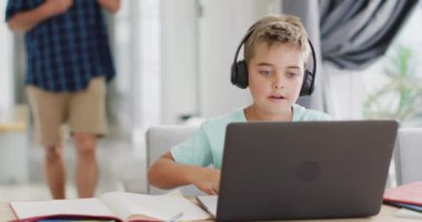 Kafkasyalı mutlu çocuk masada oturuyor ve bilgisayarını online ders olarak kullanıyor, ağır çekimde. Kaliteli zaman geçirme, ev hayatı ve çocukluk.