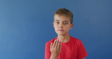 Mavi arka planda işaret dili kullanan mutlu beyaz çocuk portresi, yavaş çekim. Sağırlık, sakatlık ve çocukluk.