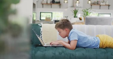 Kanepede yatıp dizüstü bilgisayar kullanan beyaz çocuk, ağır çekimde. Kaliteli zaman geçirme, ev hayatı ve çocukluk.