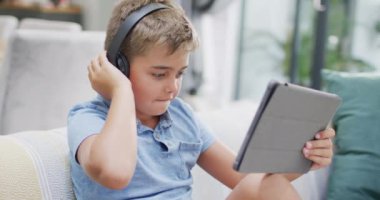 Kanepede oturup tablet kullanan mutlu beyaz çocuk, ağır çekimde. Kaliteli zaman geçirme, ev hayatı ve çocukluk.