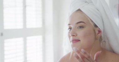 Kafkasyalı mutlu ve bornozlu kadın yüzüne kremi yavaş çekimde sürüyor. Yaşam tarzı, evde kaliteli zaman geçirme ve vücut kapsamı.