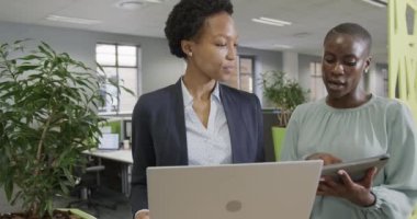 İki Afro-Amerikalı iş kadını ağır çekimde laptoplarını ve tabletlerini kullanarak işi tartışıyor. İş, takım çalışması, kapsayıcılık ve iletişim.