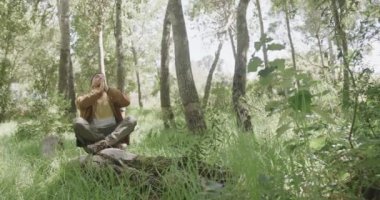 Afrika kökenli Amerikalı bir adam ağaç gövdesinde oturuyor ve ormanda meditasyon yapıyor, ağır çekimde. Kaliteli zaman geçirmek, yaşam tarzı, doğa, yürüyüş ve kamp..