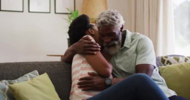 Mutlu Afro-Amerikan çifti kanepede oturup kucaklaşıyor ve gülümsüyor. Karantina süresince evde tecrit altında kalmak.