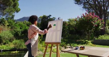 Gülümseyen Afrikalı Amerikalı kadın güneşli bahçede tuvale resim çiziyor. Karantina süresince evde tecrit altında kalmak.