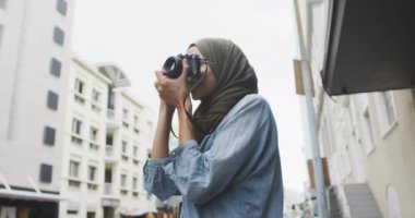 Güneşli bir günde şehirde tesettürlü giyinen, ayakta duran ve fotoğraf çeken melez bir kadının yan görüntüsü.