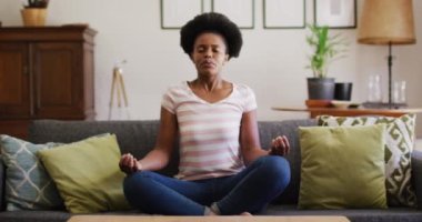 Kanepede oturmuş yoga pozu veren Afro-Amerikalı meditasyon yapıyor. Karantina süresince evde tecrit altında kalmak.