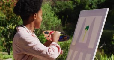 Gülümseyen Afrikalı Amerikalı kadın güneşli bahçede tuvale resim çiziyor. Karantina süresince evde tecrit altında kalmak.