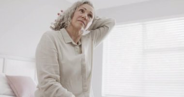 Son sınıf beyaz kadın yatakta oturuyor ve boynuna masaj yapıyor, ağır çekimde. Emeklilik ve kıdemli yaşam tarzı, model değiştirilmemiş.