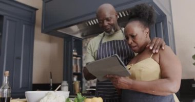 Happy senior african american couple cooking and using tablet in kitchen in slow motion. Spending quality time at home, technology, retirement and food.