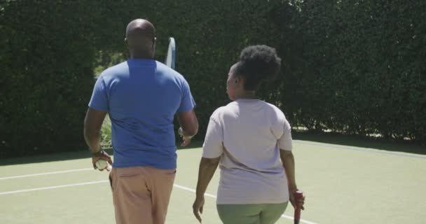テニスコートでテニスラケットを持ってゆっくりと歩くアフリカ系アメリカ人の幸せなシニアカップル 積極的なライフスタイルとスポーツ — ストック動画