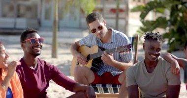 Palmiye ağaçları ve sahil evi olan plajda oturup gitar çalan mutlu bir arkadaş grubu. Tatil, boş zaman, yaz ve arkadaşlık.