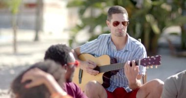 Palmiye ağaçları ve sahil evi olan plajda oturup gitar çalan mutlu bir arkadaş grubu. Tatil, boş zaman, yaz ve arkadaşlık.