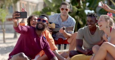 Mutlu bir grup arkadaş selfie çekiyor ve palmiye ağaçlarıyla sahilde gitar çalıyorlar. Tatil, boş zaman, teknoloji ve arkadaşlık.