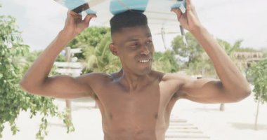 Güneşli sahilde sörf tahtası taşıyan gülümseyen Afro-Amerikan bir adamın portresi, yavaş çekim. Yaz, sağlıklı yaşam tarzı, hobiler, rahatlama ve tatiller.