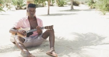 Mutlu Afro-Amerikalı adam ağacın altında oturmuş gitar çalıyor ve güneşli sahilde yavaş çekimde şarkı söylüyor. Yaz, hobiler, rahatlama ve tatiller.