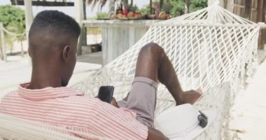Afrika kökenli Amerikalı adam hamakta güneşli sahilde akıllı telefon kullanarak uzanıyor, ağır çekimde. Yaz, rahatlama, iletişim ve tatiller.