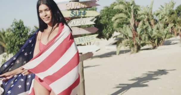日当たりの良いビーチで看板によってアメリカの国旗で踊る幸せなヒスパニック系の女性 スローモーション リラックスと休暇 — ストック動画
