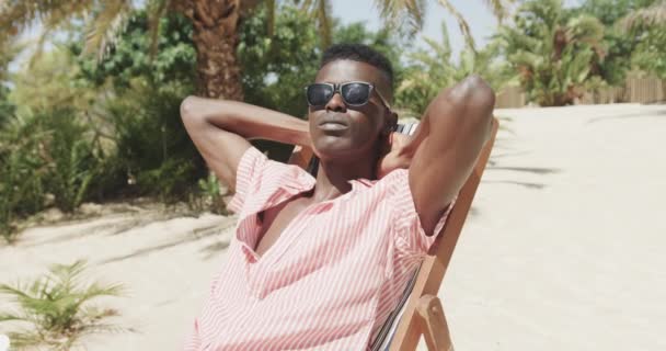 Afrykański Amerykanin Siedzący Leżaku Okularach Przeciwsłonecznych Relaksujący Się Słonecznej Plaży — Wideo stockowe