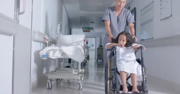 病院の廊下で車椅子に座っている多様な男性看護師と子供の患者 スローモーションで — ストック動画