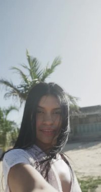 Güneşli sahilde gülümseyen mutlu, melez bir kadının dikey video portresi, yavaş çekim. Yaz, rahatlama, iletişim ve tatiller.